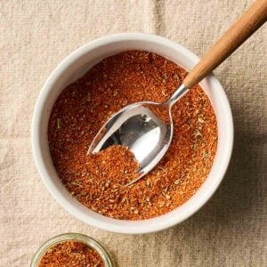 A bowl with a spice mix inside and a spoon, with a smaller jar to the side.
