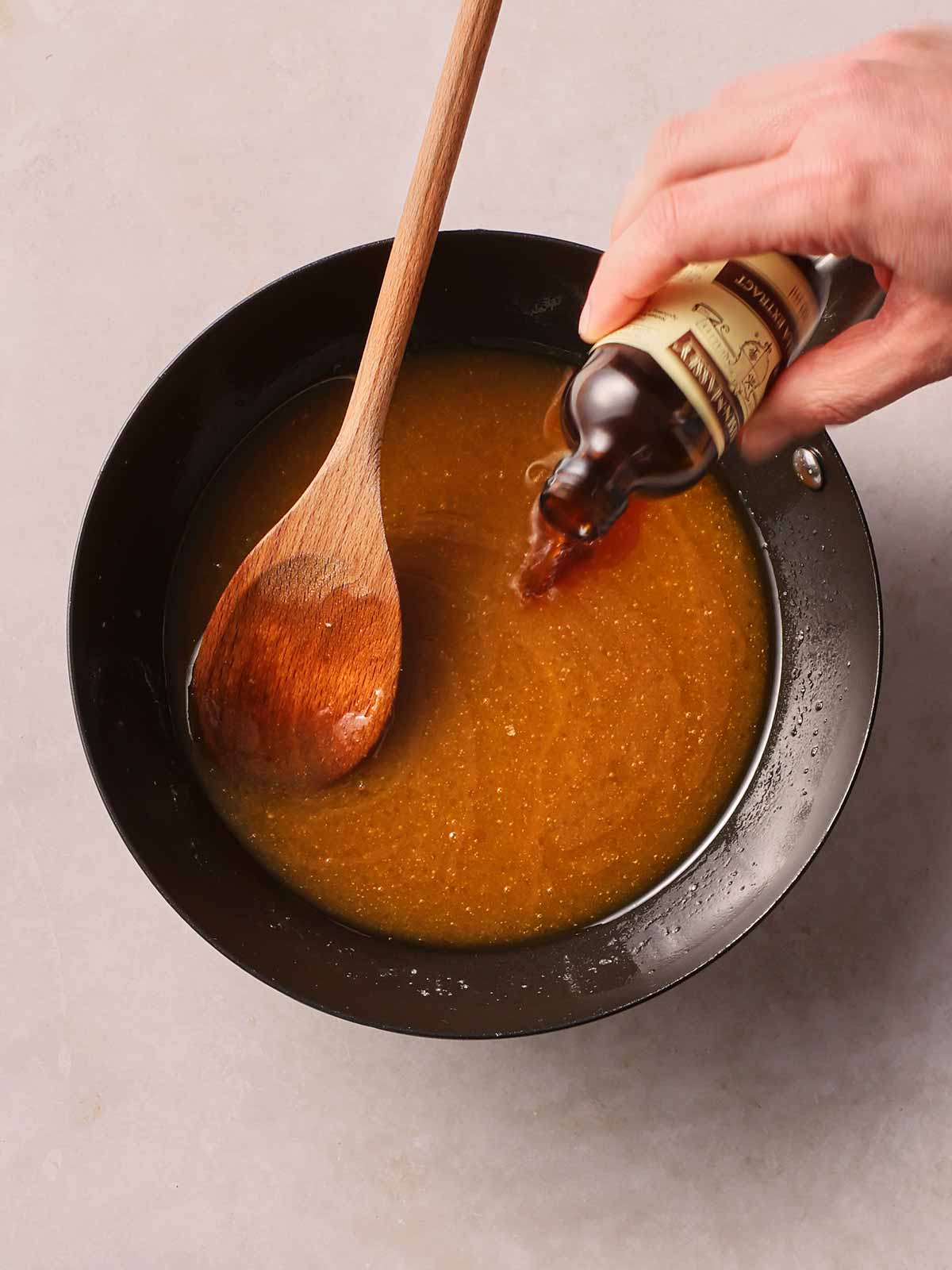 A pan with melted brown liquid in with a wooden spoon, with a hand pouring in vanilla extract.