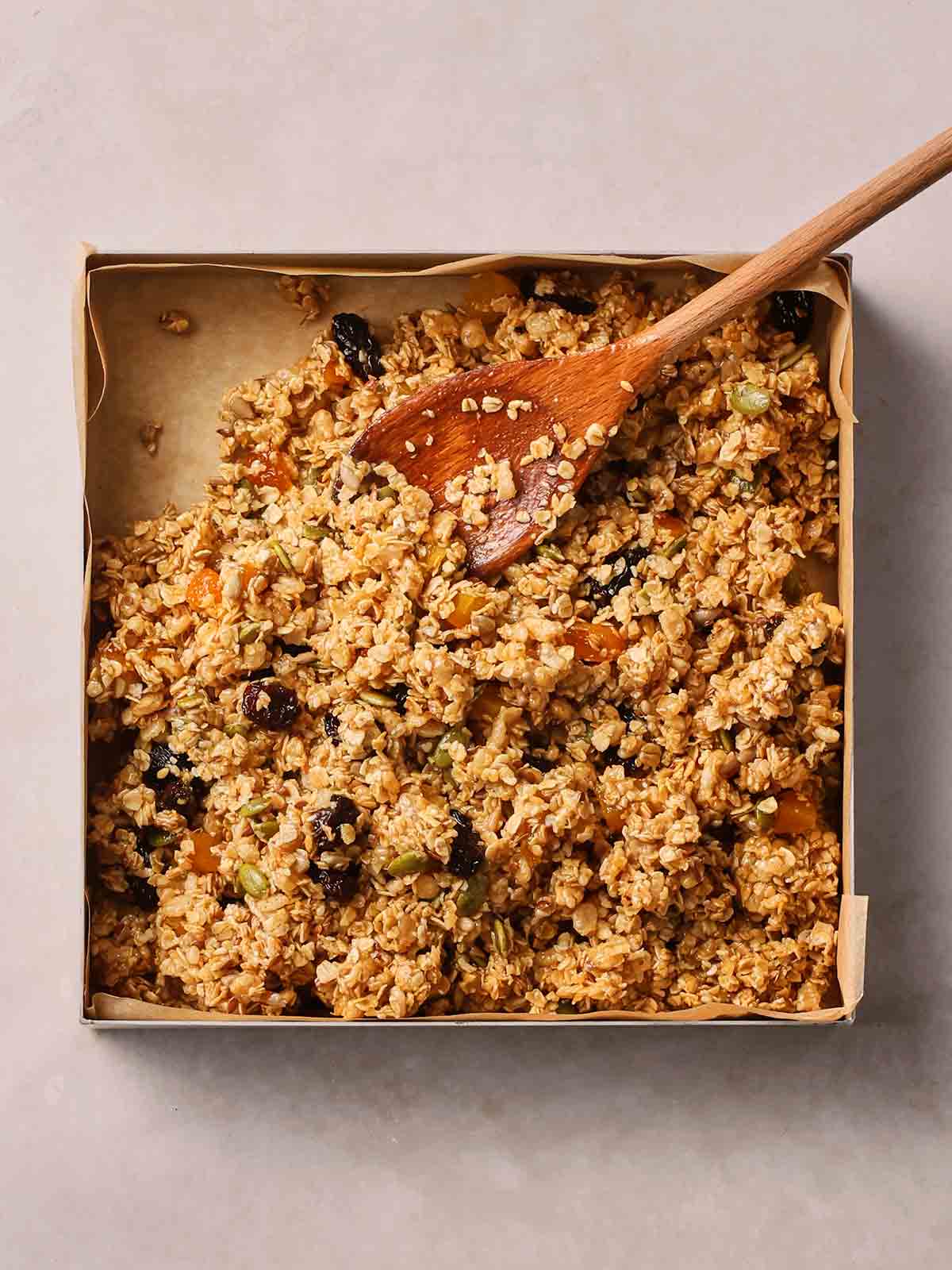 A wet oats mixture with seeds and fruit having been poured into a lined, square tin, with a wooden spoon in.