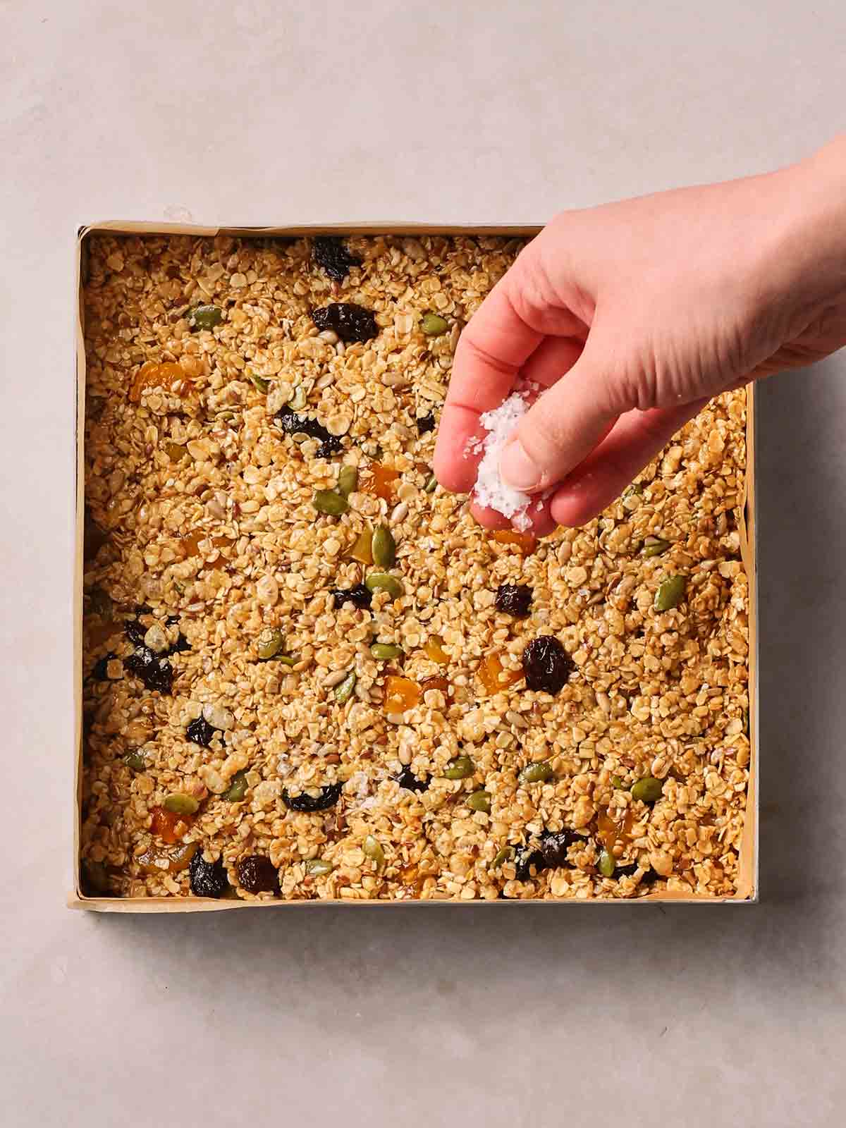 A square tin on a white surface with an oat mixture with seeds and nuts in, and a hand sprinkling over sea salt, before the granola bars are baked.
