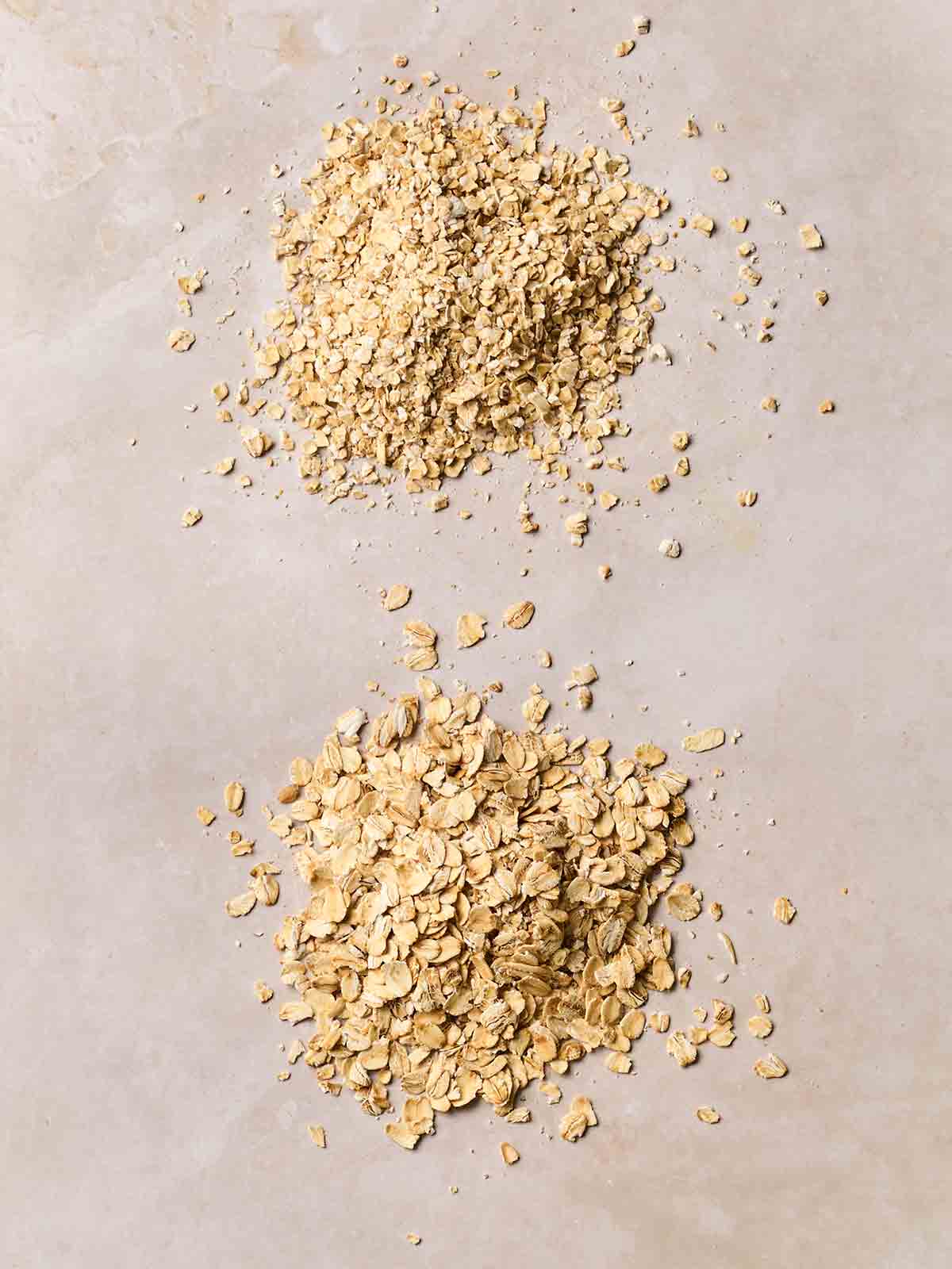 Two piles of different style oats on a white counter. One at the top is quick oats, the bottom pile is jumbo oats.