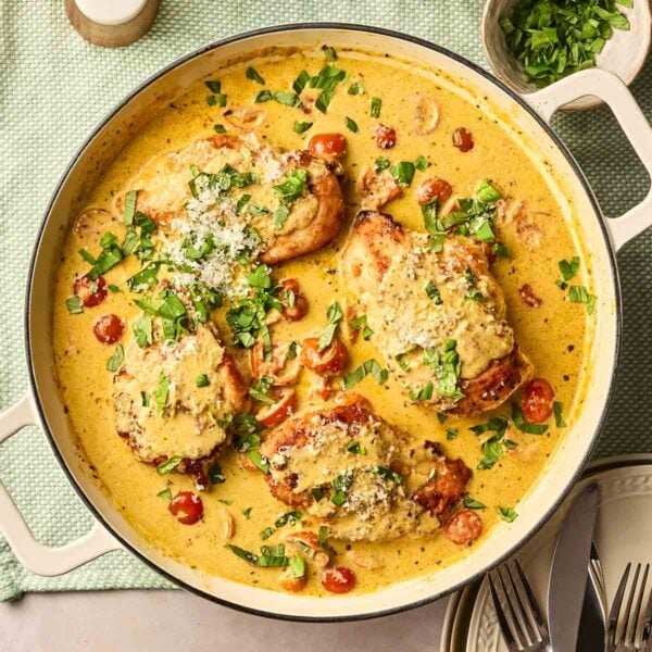 A large pan of chicken in a creamy pesto sauce, with tomatoes and basil, ready to serve onto plates on a dining table.