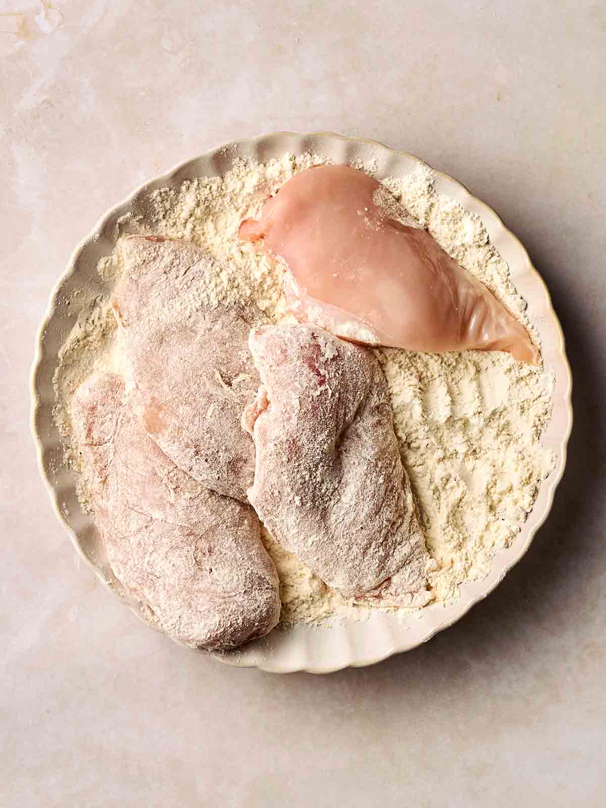 Four chicken breasts being coated in flour on a white plate.