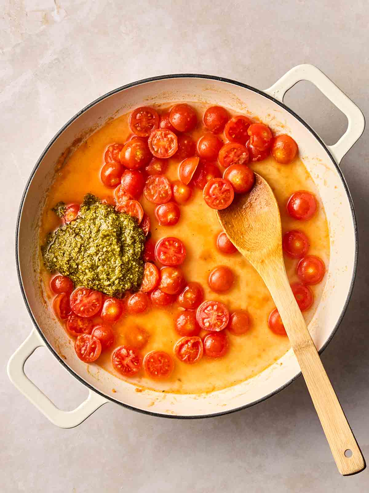 A pan filled with chicken stock, halved cherry tomatoes, a dollop of pesto and a wooden spoon.