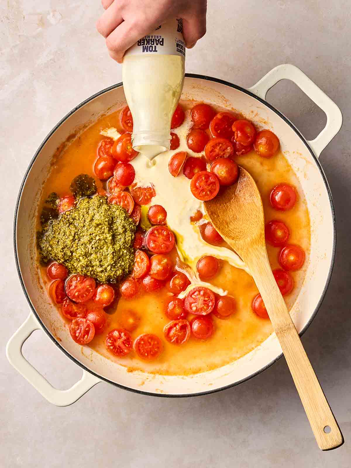 A hand pouring some double cream into a pan of stock, tomatoes and pesto.