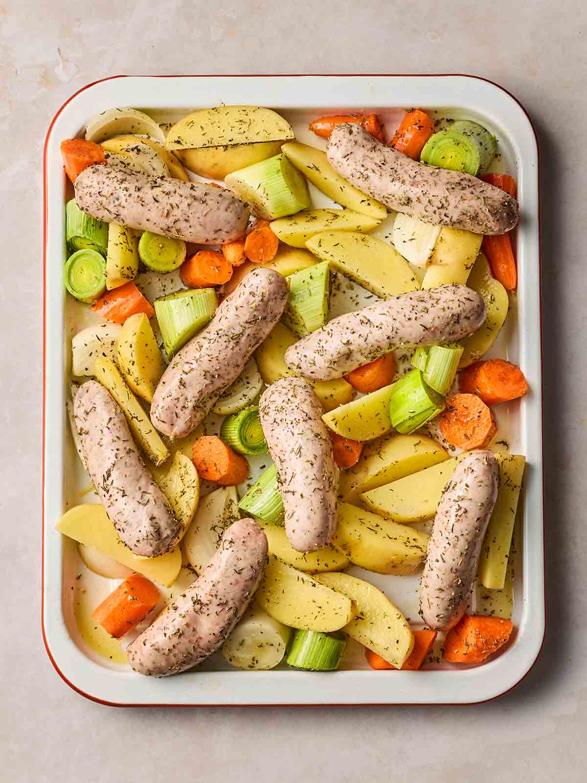 An oven tray filled with with casserole vegetables and chunky sausages with herbs sprinkled over, ready to go in the oven.