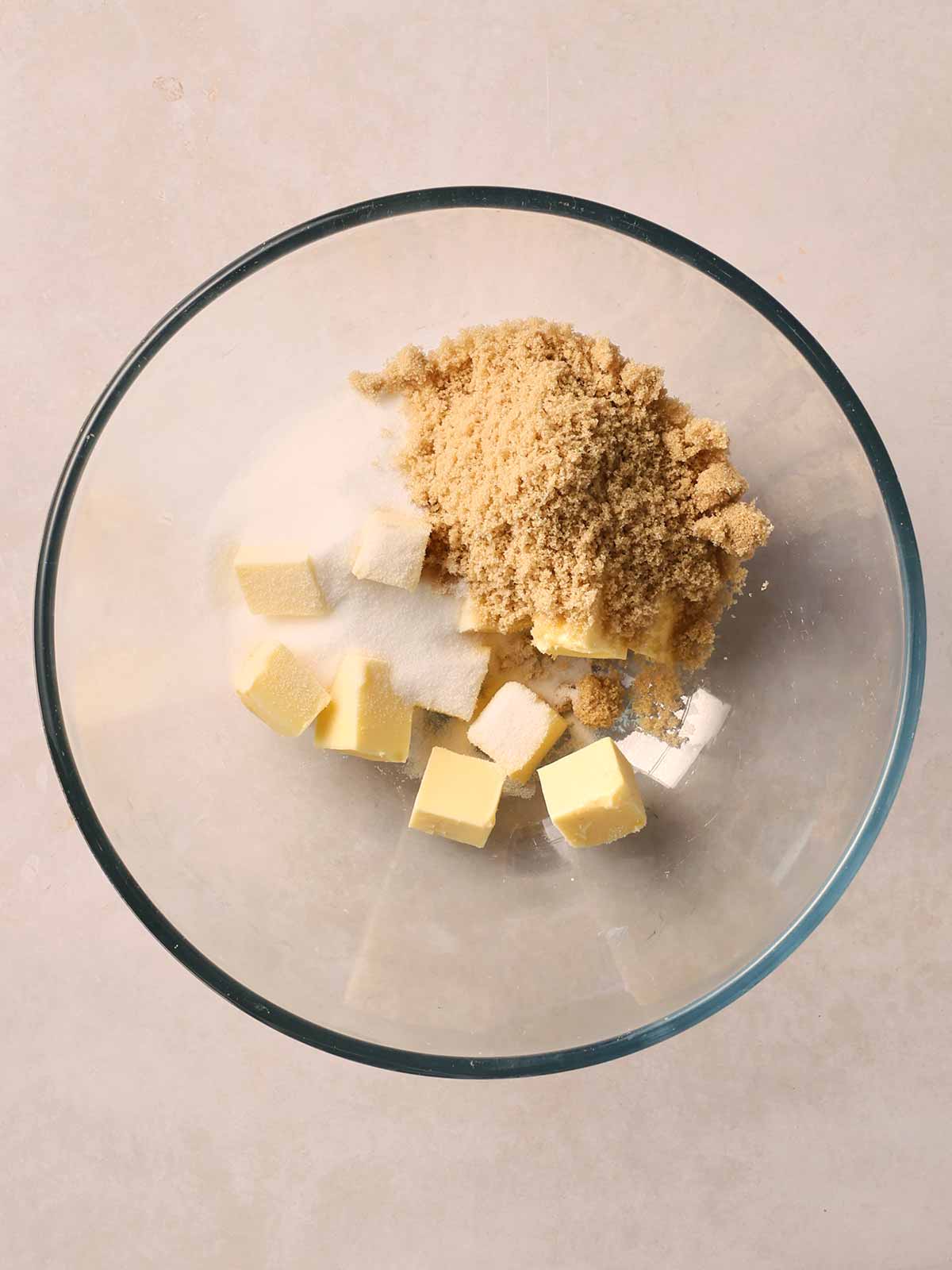 A glass bowl with cubes of butter, caster sugar and light brown sugar inside, on a white counter.