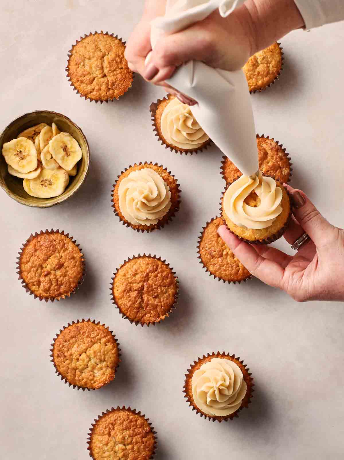Two hands over a batch of banana cupcakes on a white surface, decorating them with buttercream with a piping bag.
