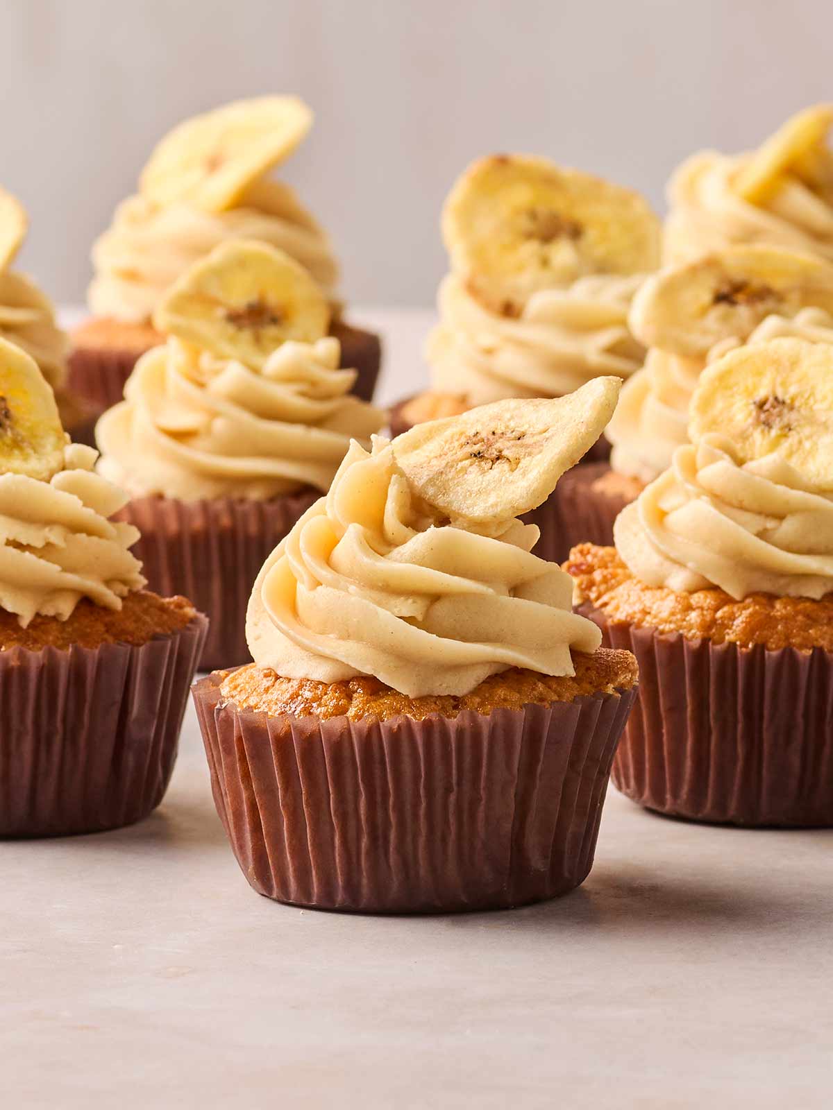 A close up of a batch of banana cupcakes, with sliced banana chips on top and buttercream frosting.