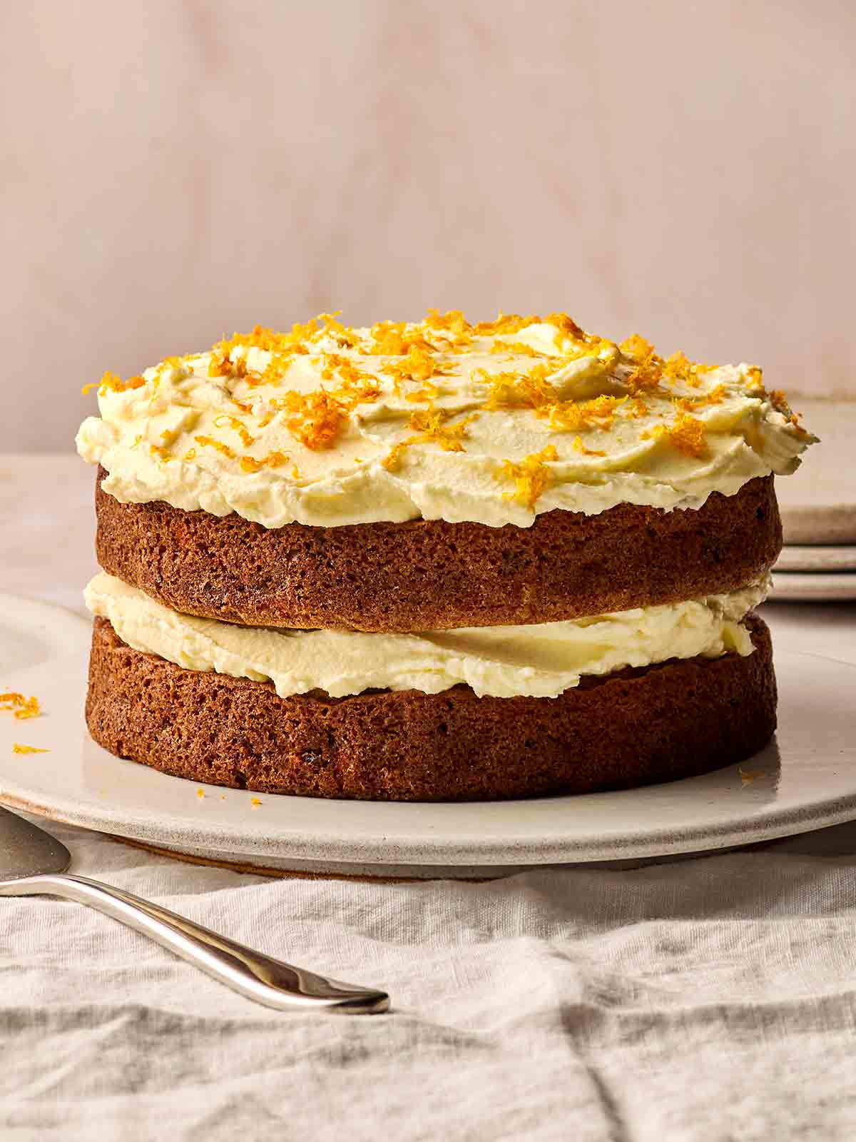 A two-tier Carrot Cake on a plate from the side, with buttercream icing in the middle and on top to decorate.