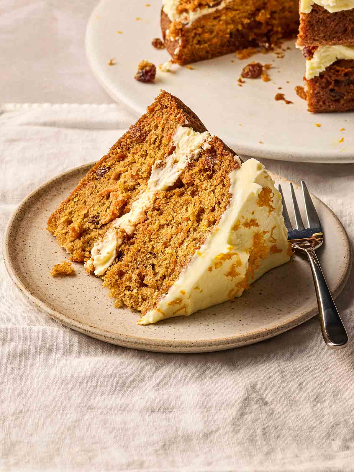 A slice of homemade Carrot Cake on a plate with a cake fork, with the rest of the cake in the background.