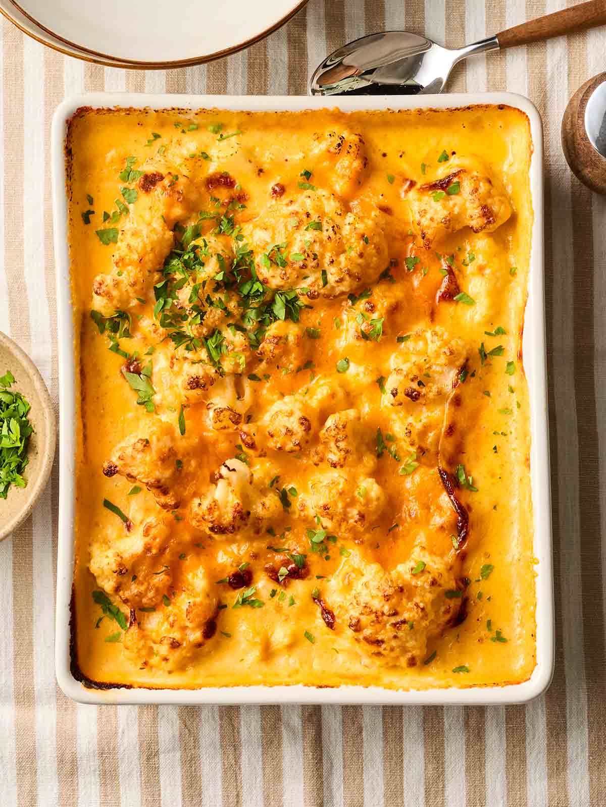 An oven dish filled with baked Cauliflower Cheese on a table with a stripy tablecloth, ready to be served.