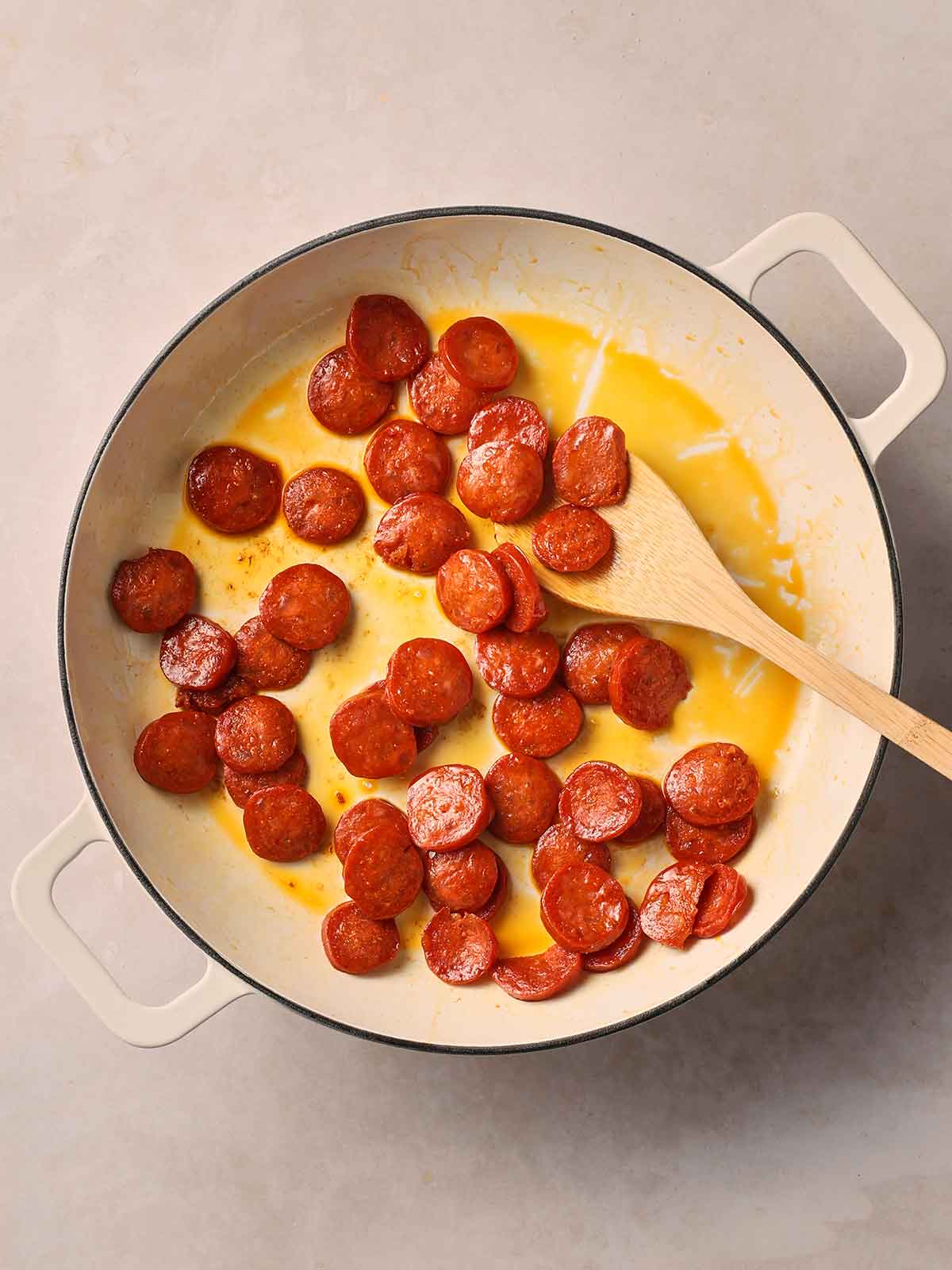 Sliced chorizo frying in a pan with the oils released, with a wooden spoon in.