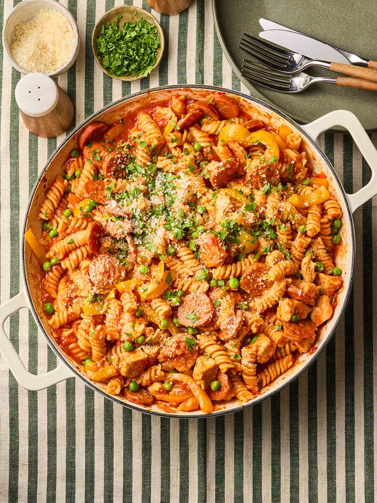 A pan of chicken and chorizo pasta with peas on a table with a stripy tablecloth, ready to be served.