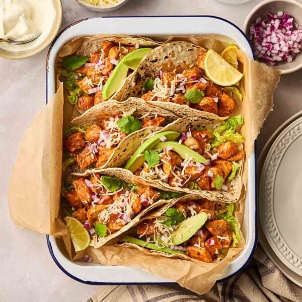 An oven dish filled with cooked and filled chicken tacos, with avocado on top and cheese, with lots of other toppings around the edges on a table.