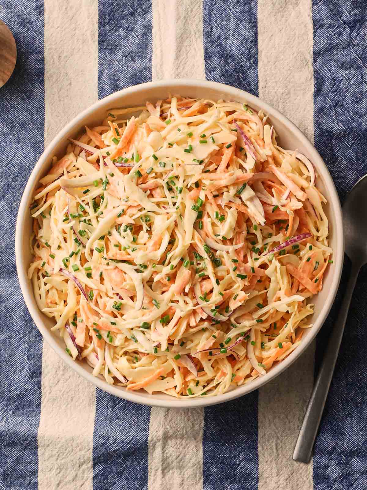 A bowl of homemade coleslaw on a blue and white stripy tablecloth, with a spoon to the side.