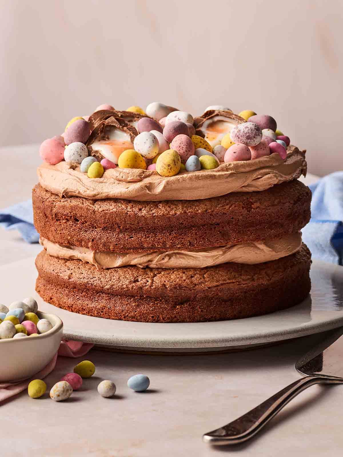 A two-tier Easter chocolate cake with buttercream icing in the middle and on top, with lots of mini eggs on top too. Ready to slice on a plate.