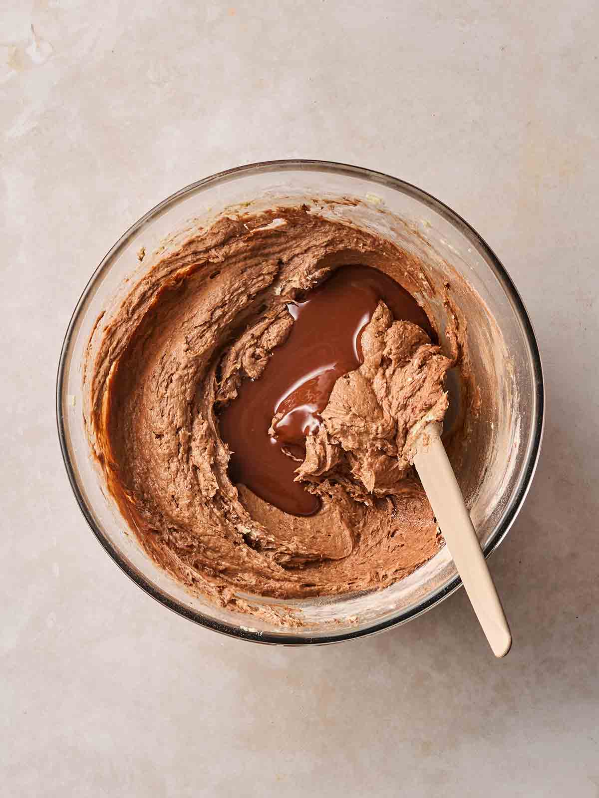 A glass bowl on a white surface, with chocolate cake batter inside, with melted chocolate ready to mix in with a wooden spoon.