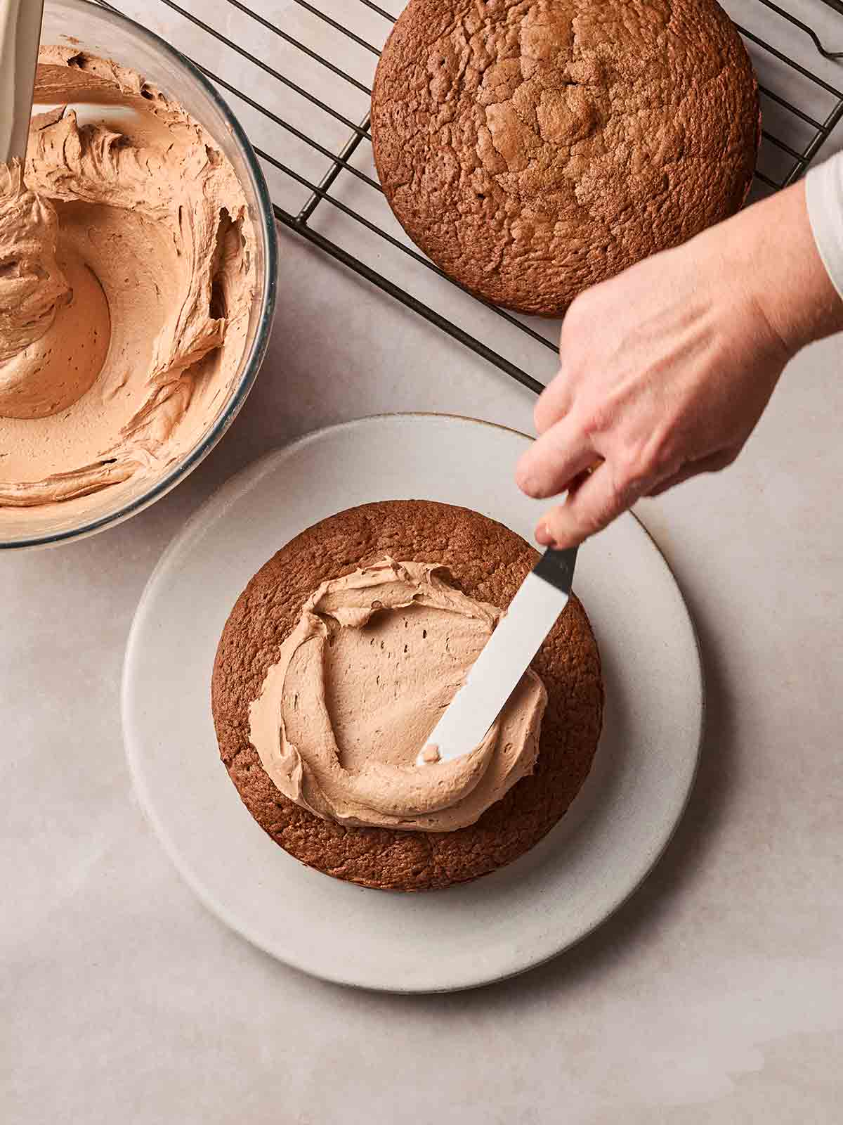 A hand adding chocolate buttercream to a chocolate sponge cake, with the cooled cake next to it.