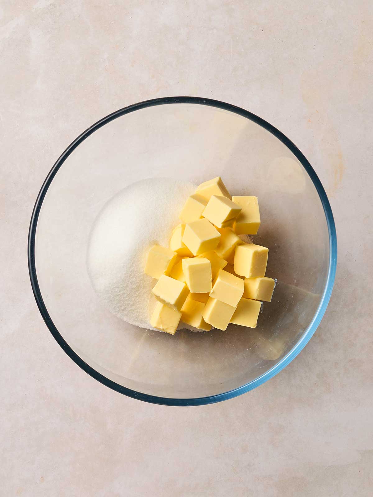 A glass bowl on white surface containing cubed butter and sugar.