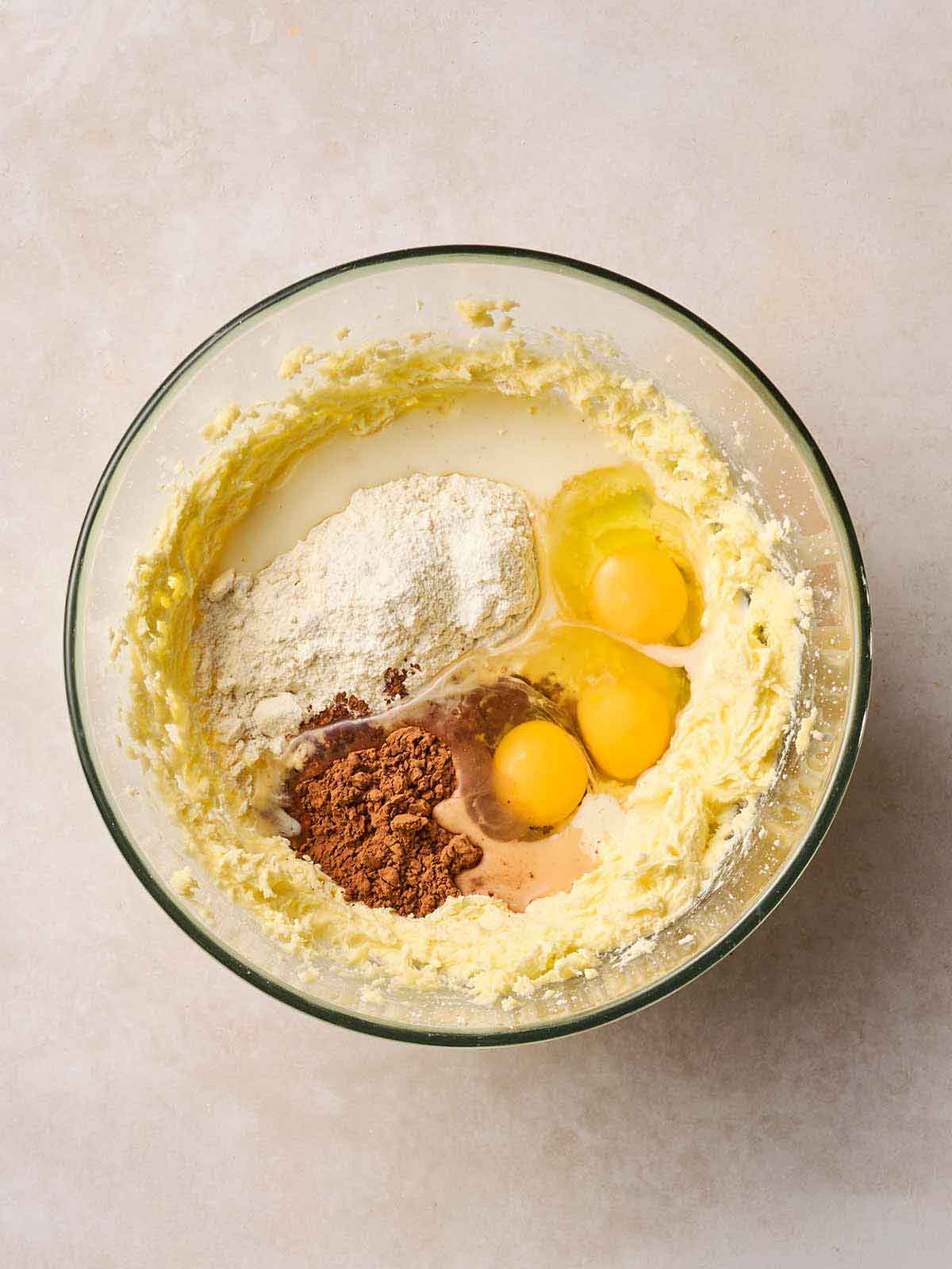 A glass bowl with cupcake batter ingredients inside, half mixed together, including eggs, creamed butter, flour and cocoa.