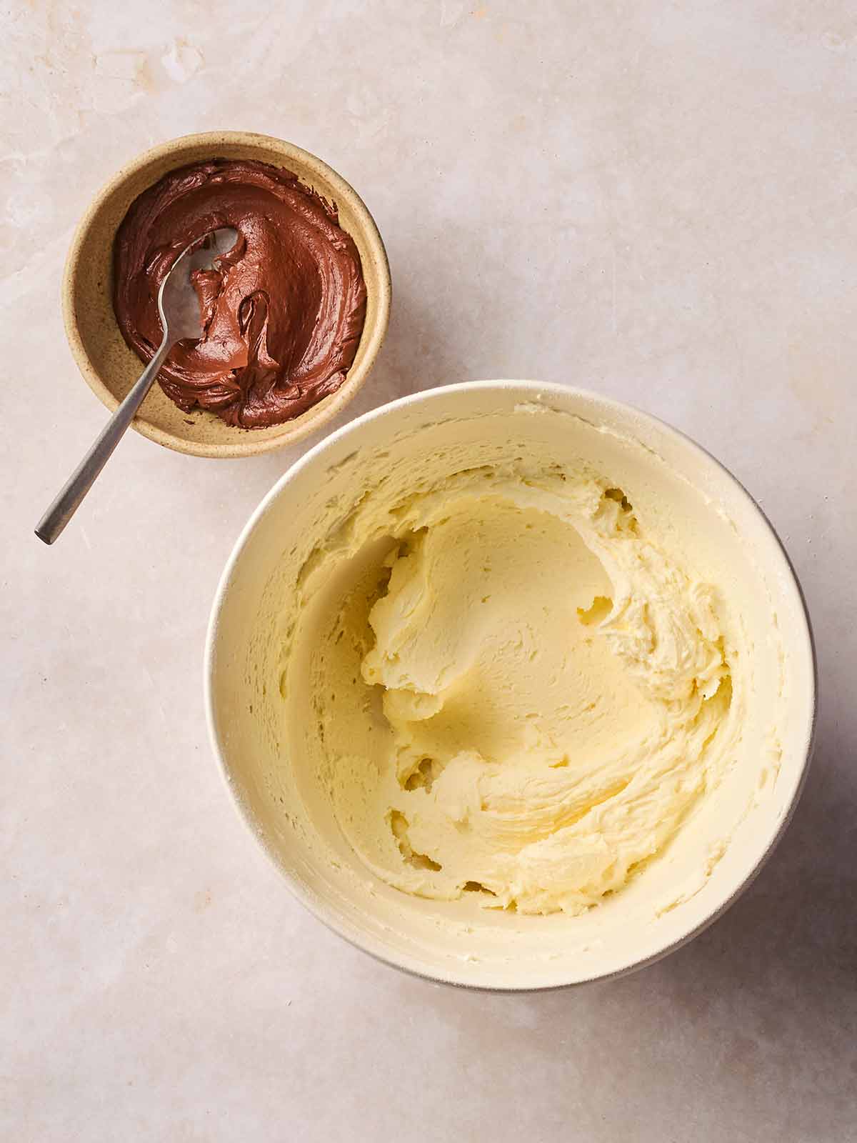 Two bowls on a white surface, one with cream frosting, another with mixed cocoa.