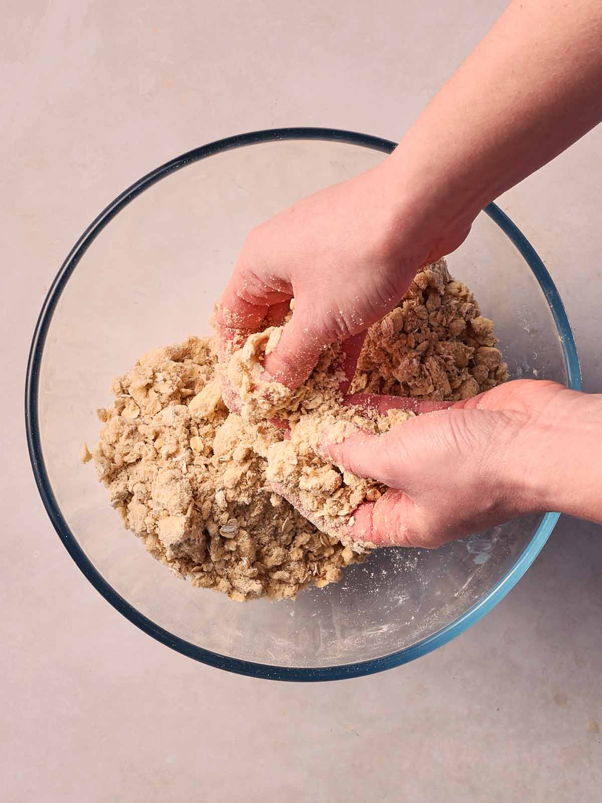 Two hands over a glass bowl on a white surface, rubbing a crumble topping.