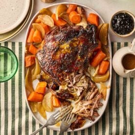 A cooked lamb shoulder joint on a platter with onions and carrots, on a set dining table, ready to be dished up.