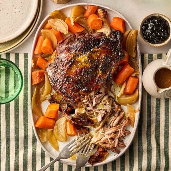 A cooked lamb shoulder joint on a platter with onions and carrots, on a set dining table, ready to be dished up.