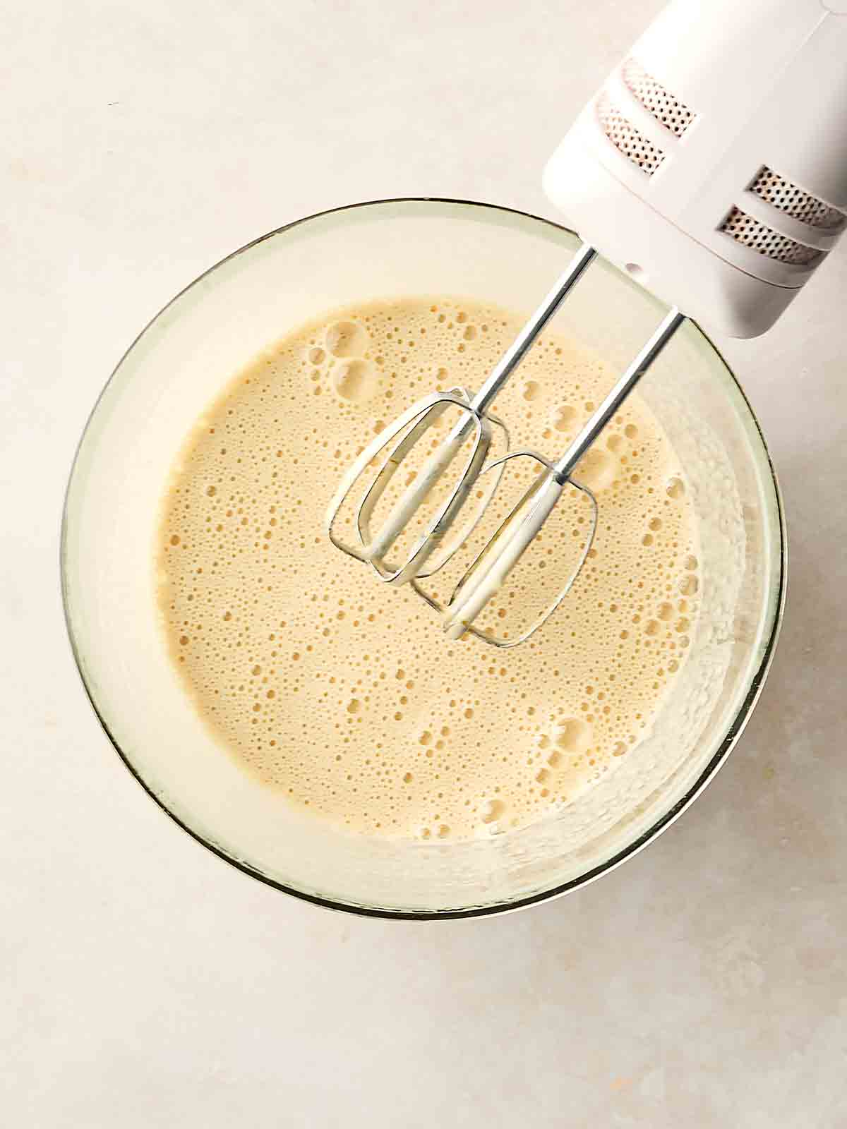 A white mixture with foamy bubbles in a glass bowl with a handheld whisk just above it.