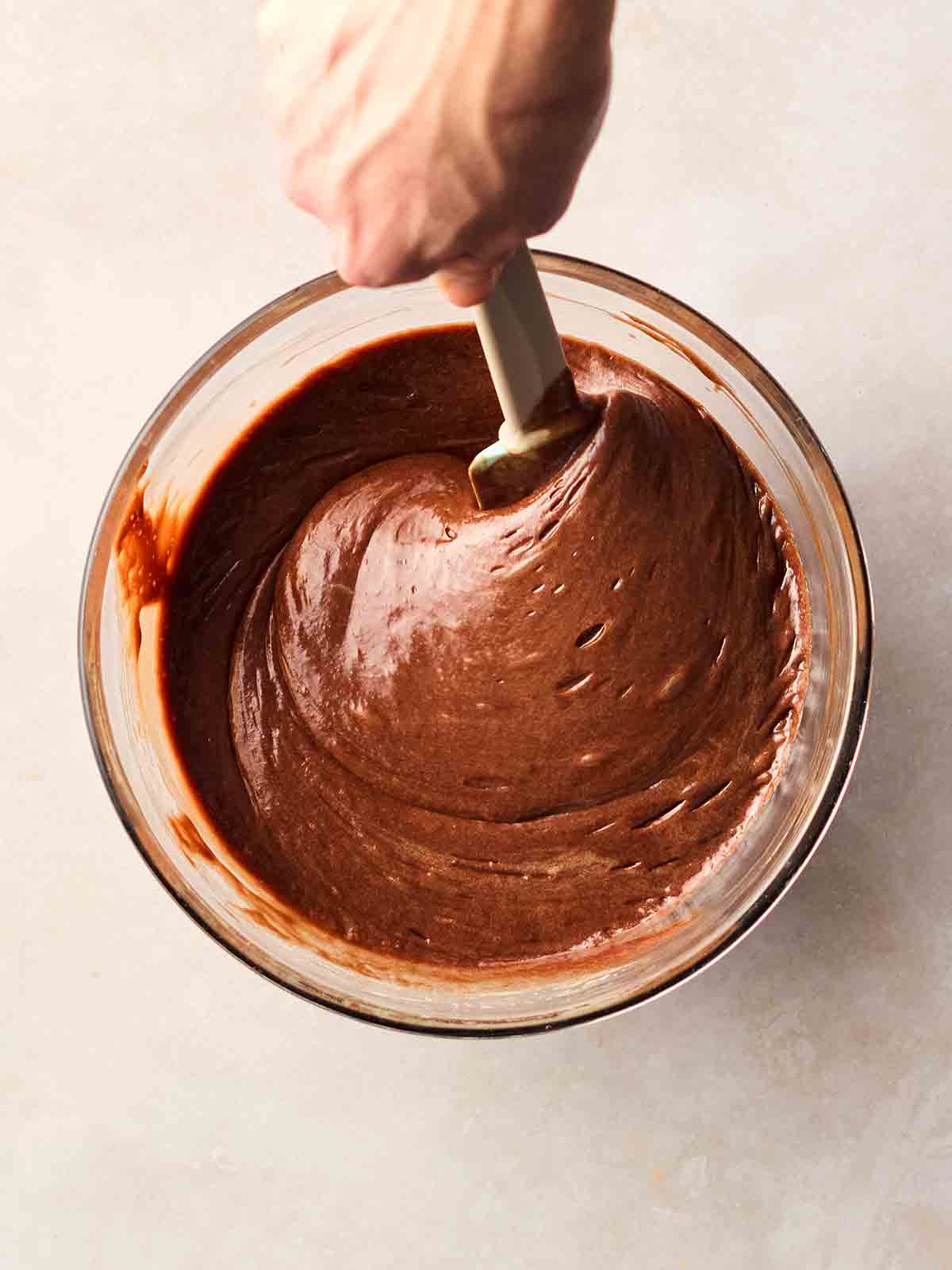 A hand stirring a bowl of melted chocolate brownie mixture on a white surface.