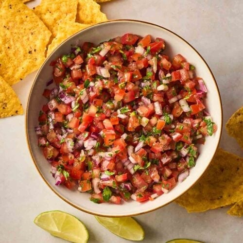 A bowl of fresh salsa with a white background with tortilla chips around it.