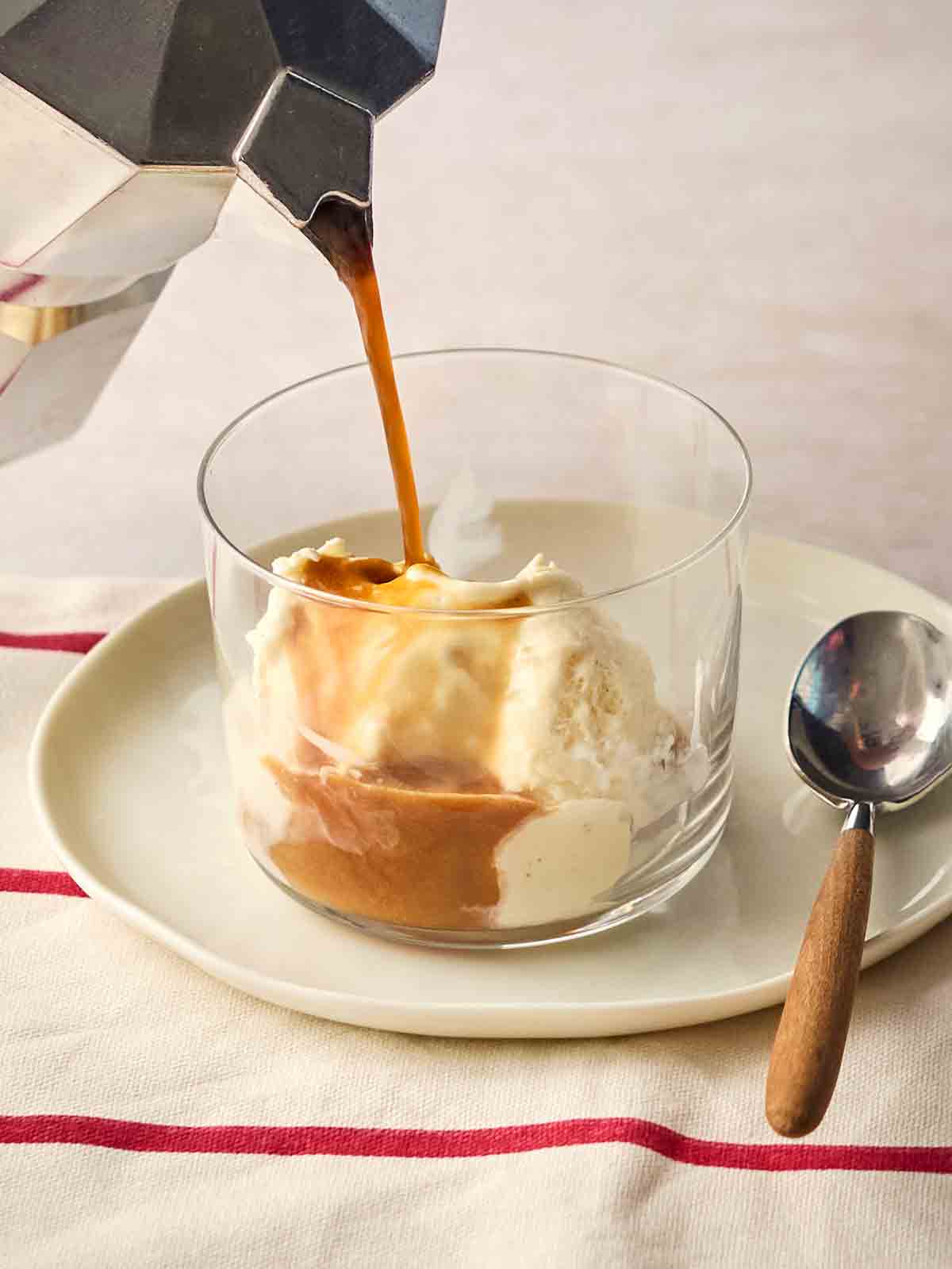 A coffee pot pouring espresso over a dessert glass with vanilla ice cream inside for an Italian Affogato dessert being served on a dining table with a spoon to the side.