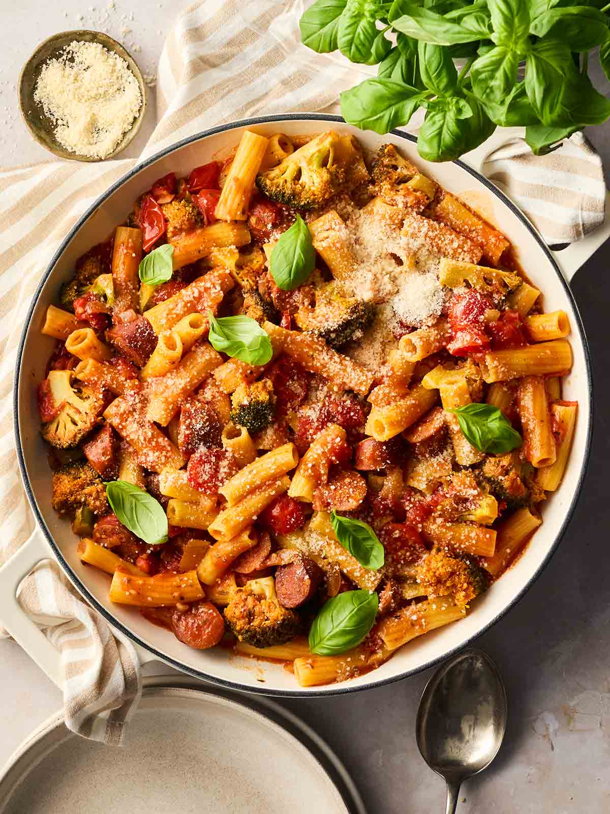 A large pan of cooked pasta with chorizo and vegetables in a tomato sauce sauce. On a table with a basil plant and sides dishes.