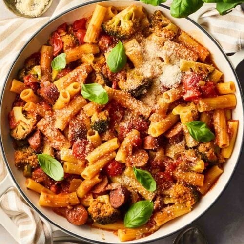 A large pan filled with chorizo pasta in a tomato sauce, on a table with dishes, a basil plant and parmesan to serve.