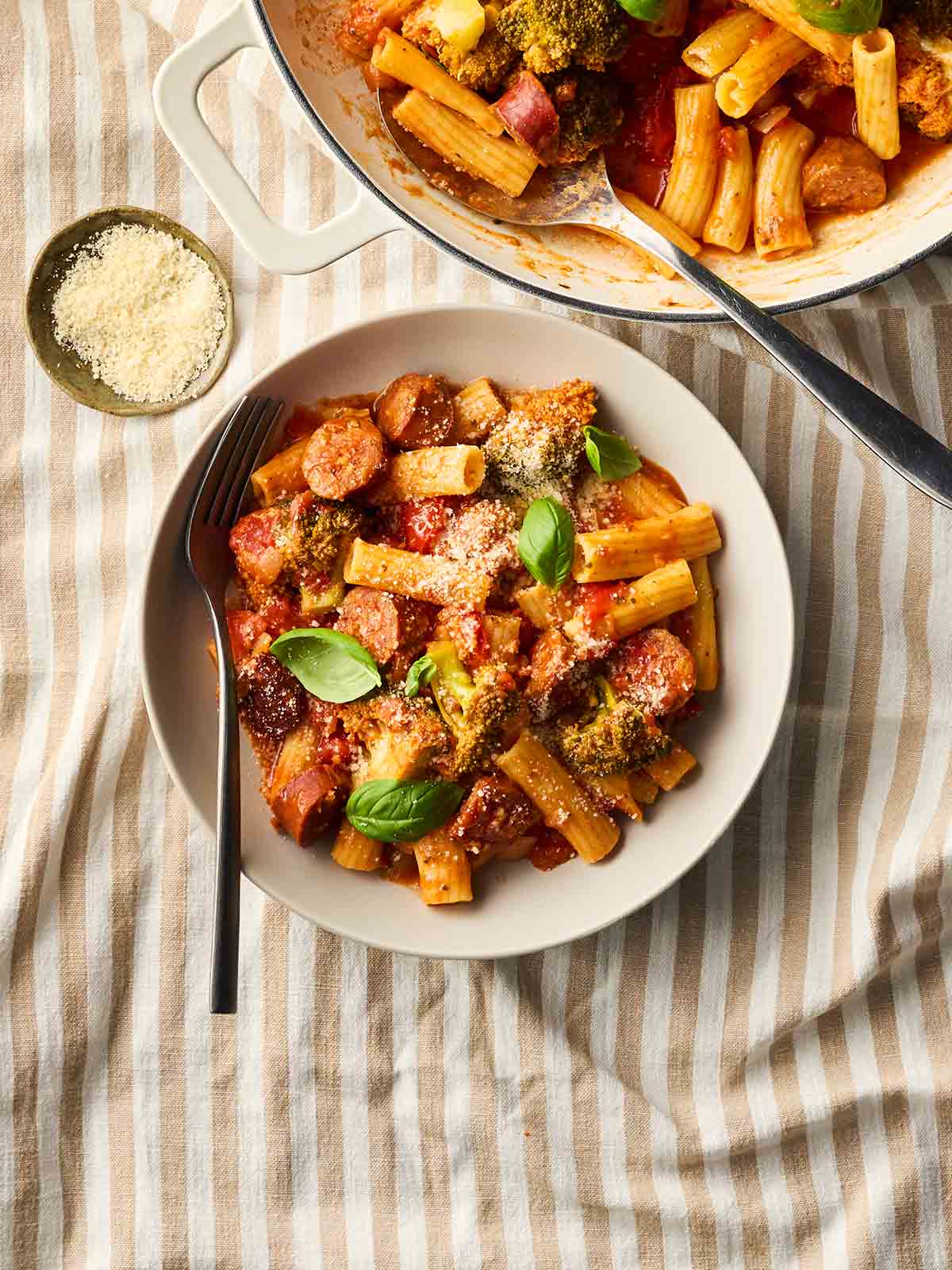 A bowl of chorizo pasta with a fork, on a tablecloth with stripes, with the pan of pasta in the corner.