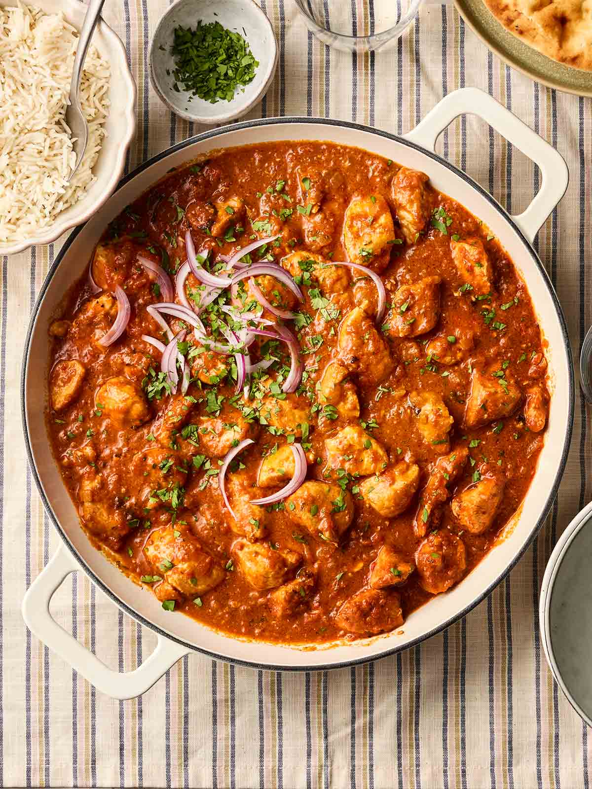 A pan in the middle of a table filled with easy chicken curry, garnished with red onion and coriander, with side dishes to the side, including rice, coriander and naan.