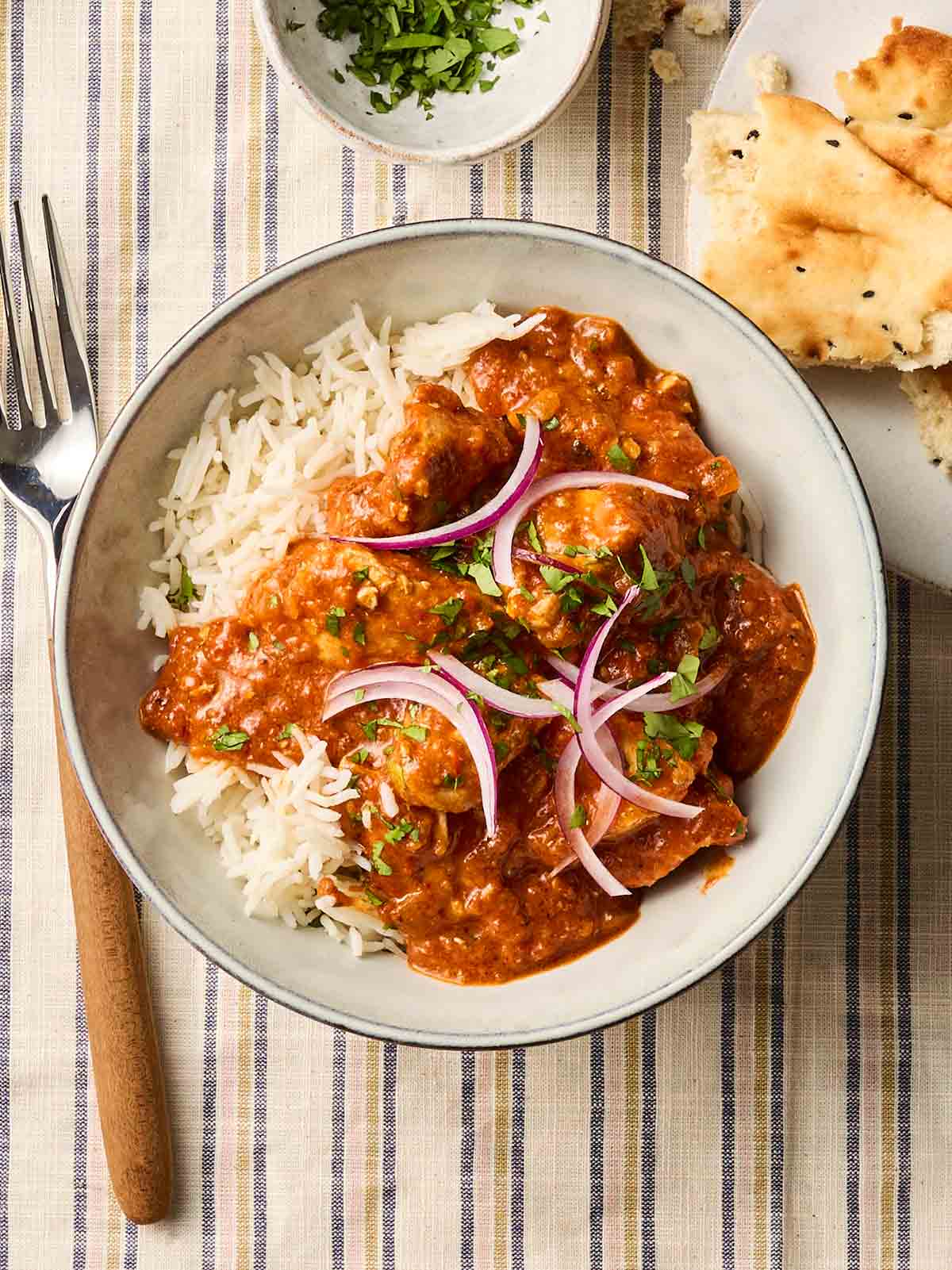 A bowl of homemade chicken curry with red onion and coriander on top, with a fork to the side and naan to serve.