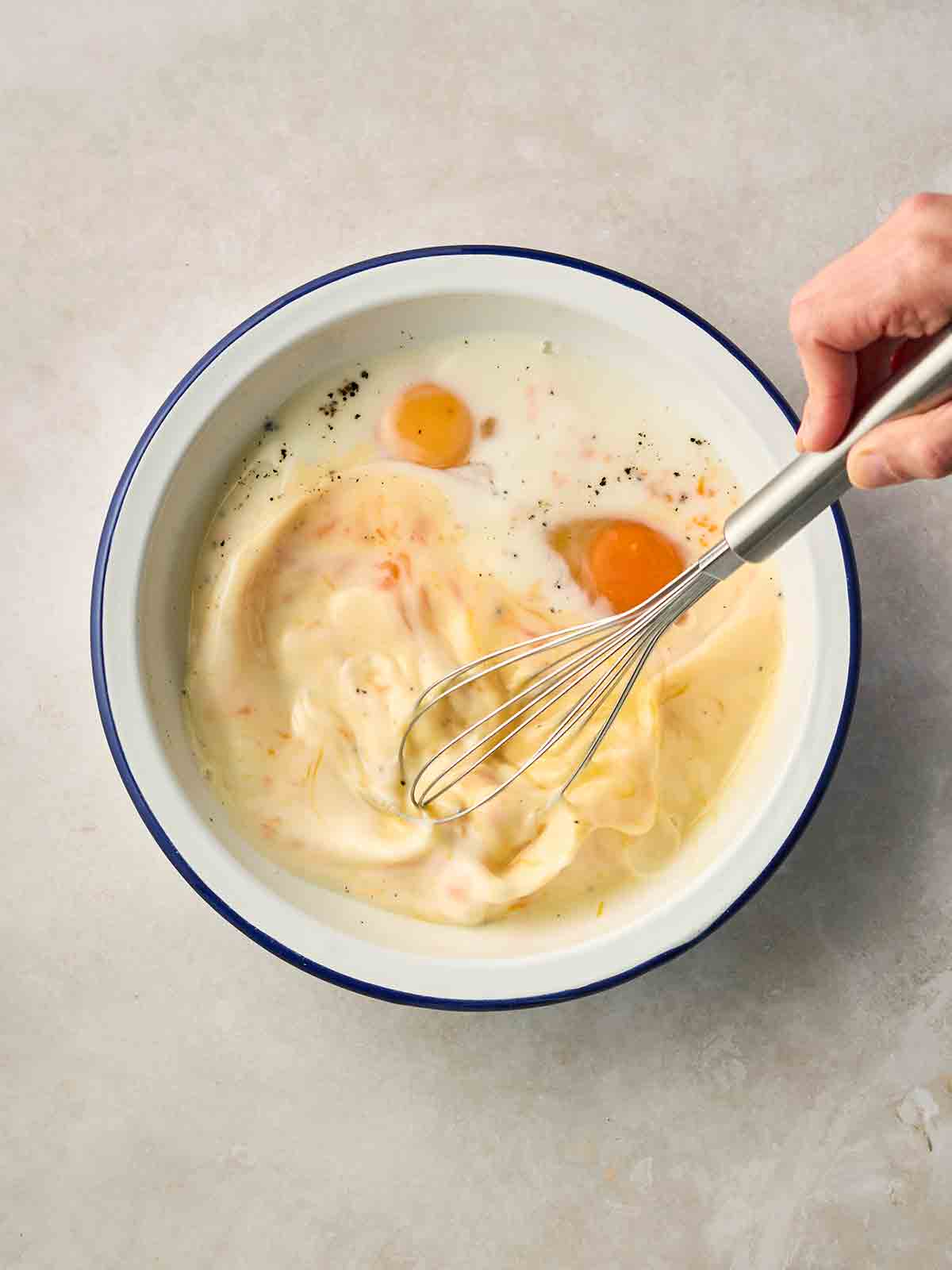 A hand holding a whisk, whisking a mixture of eggs and milk in a wide bowl.