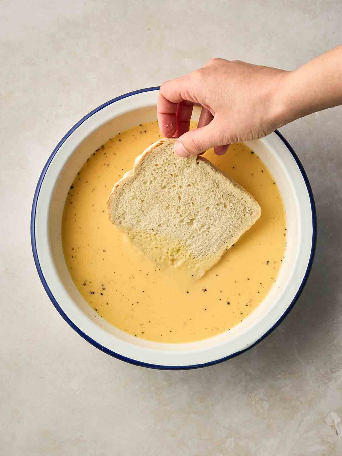 A hand placing a slice of white bread into an eggy mixture.