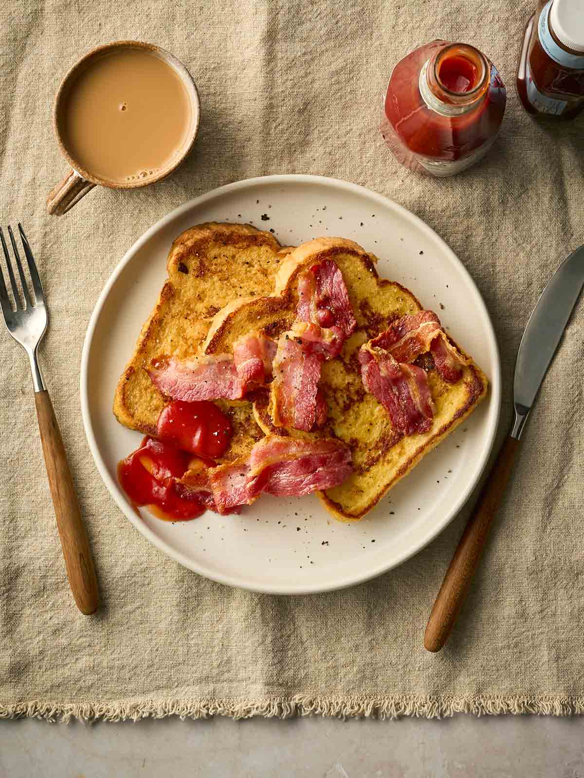 Eggy Bread with bacon at a place setting on a table with a cup of tea and ketchup to the side.