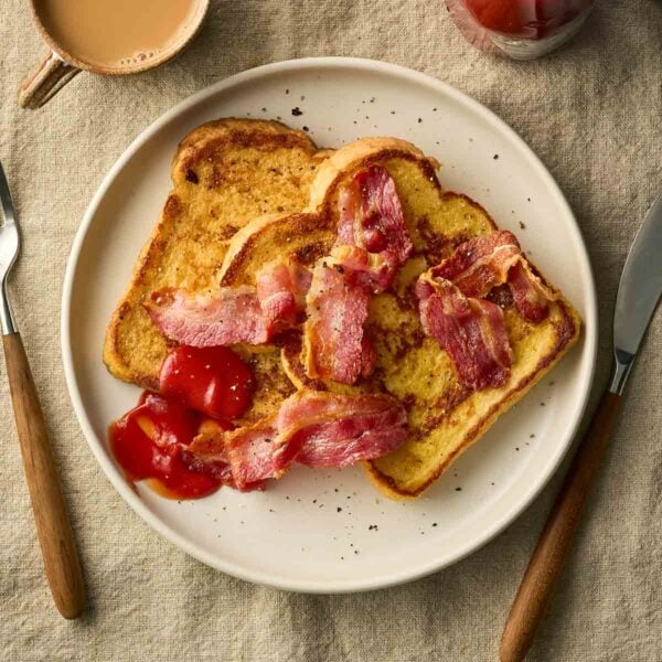 Two slices of white bread on a place with bacon on top, with cutlery to the side and a cup of tea.