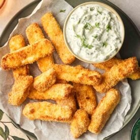 A close up of a plate of golden, crispy halloumi fries, with a bowl of homemade tzatziki to the side for dipping.