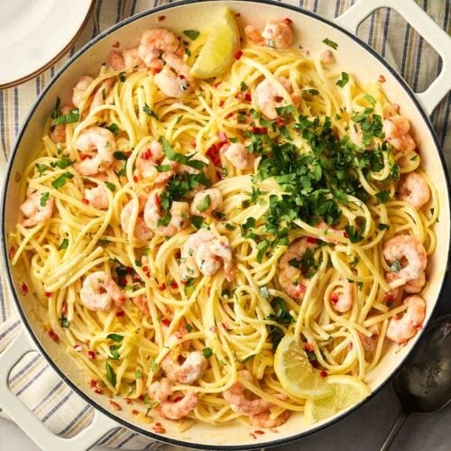 A pan of prawn linguine with two handles, in the middle of a table with a stripy cloth underneath, a spoon to these side, as well as a glass and salt and pepper. A plate is in the corner.