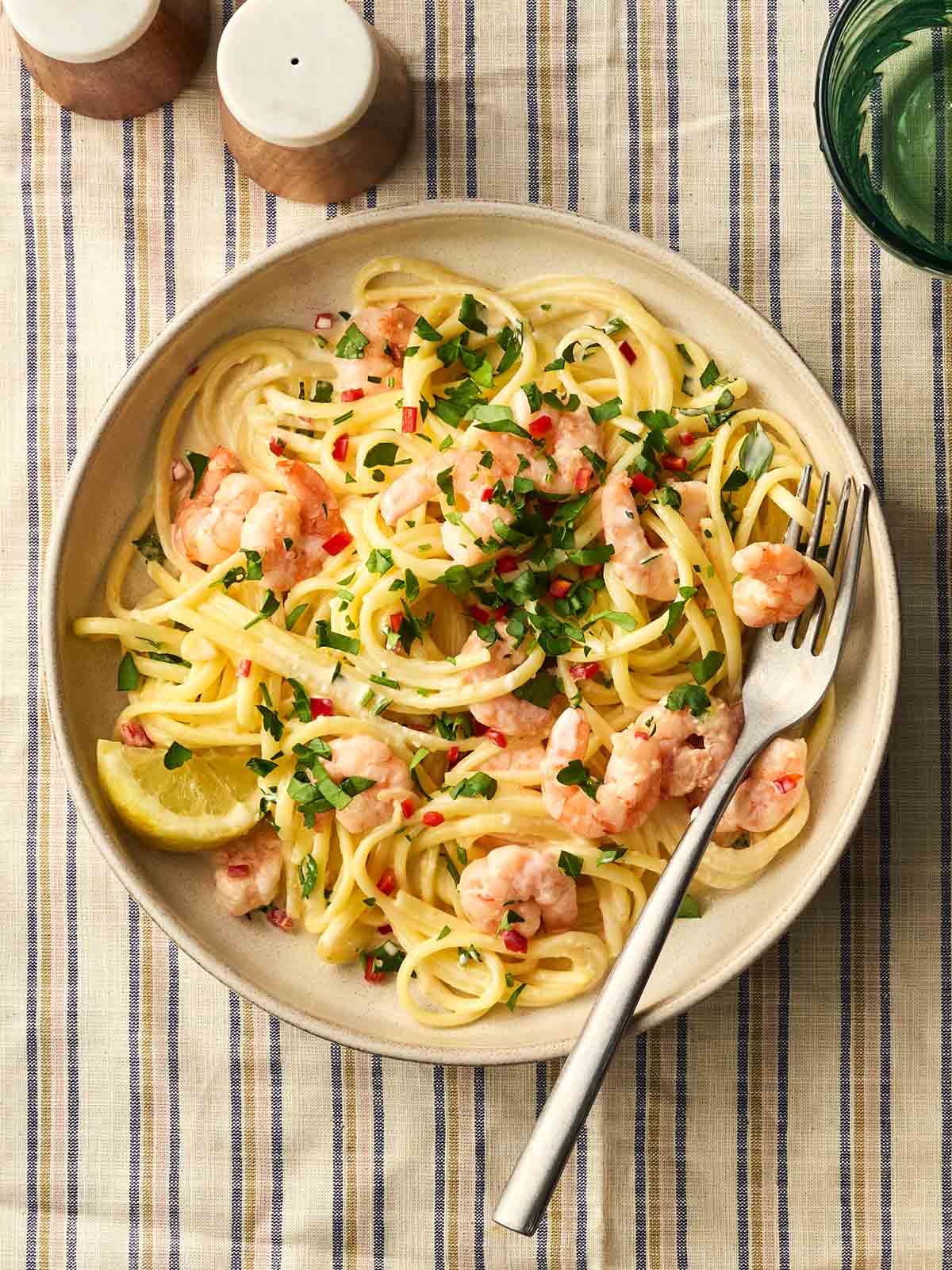 A plate of shrimp linguine with a fork in, ready to be be eaten, on a stripy tablecloth with salt and pepper to the side.