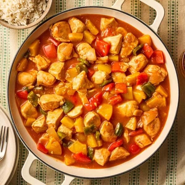 A large pot of Sweet and Sour Chicken on a green stripy tablecloth, with rice to the side, and plates and cutlery to the other side.
