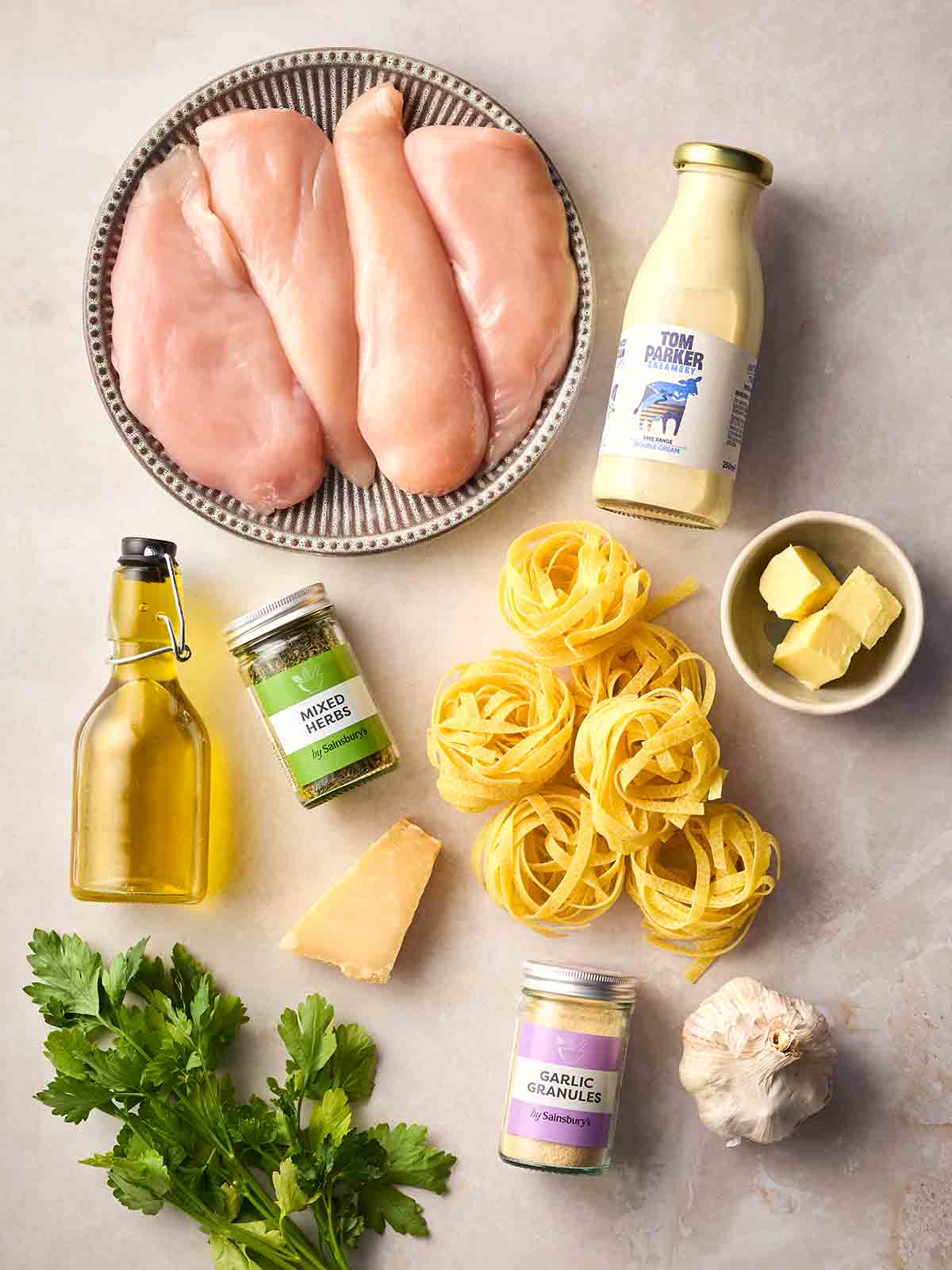Raw ingredients for making Chicken Alfredo laid out on a white counter, including chicken breasts, tagliatelle, cream, oil, butter, parmesan and herbs.