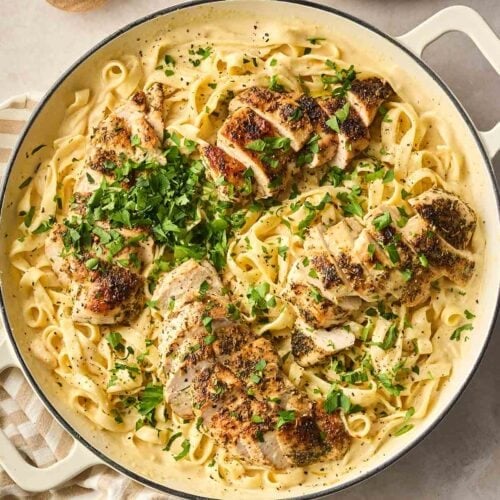 A large bowl of pasta with chicken on a counter, with pepper to the side and bowls to dish into, and a stripy dish towel.