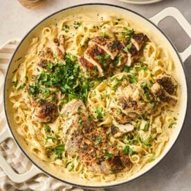 A close up of a large white pan filled with Chicken Alfredo, a dish with long tagliatelle pasta, creamy sauce and sliced herby chicken breast on top, with parsley garnished.