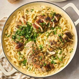 A close up of a large white pan filled with Chicken Alfredo, a dish with long tagliatelle pasta, creamy sauce and sliced herby chicken breast on top, with parsley garnished.